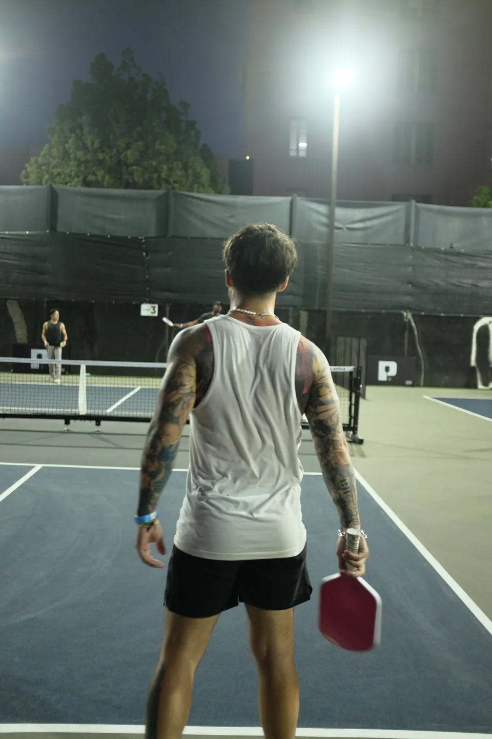 Tattooed player in a white tank top holds a red paddle at the baseline, facing his opponent across a night court