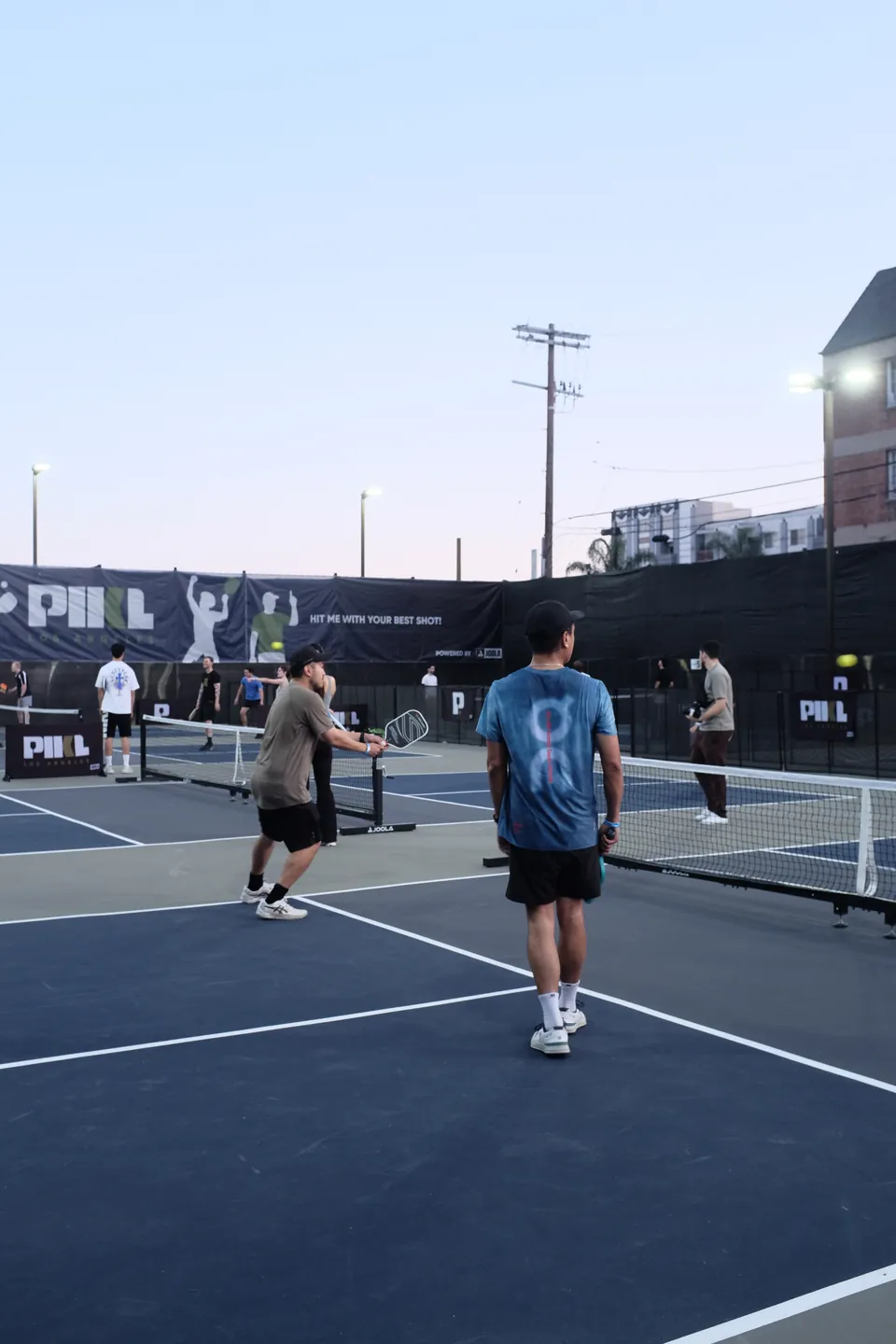 Doubles on the PIKL courts at dusk, a player in a blue tee walking the baseline in front of a PIKL banner