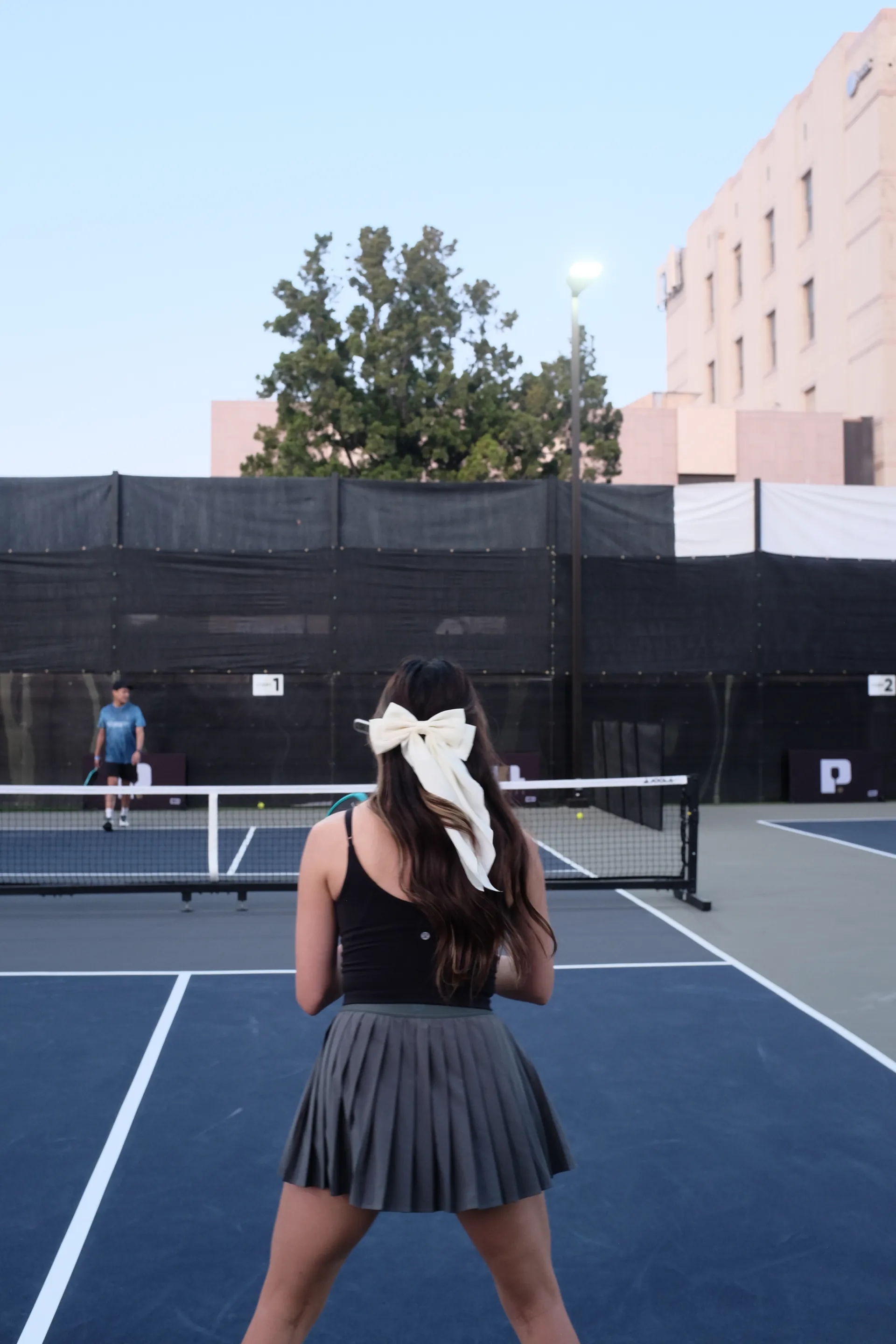 Woman with a cream hair bow and pleated grey skirt waits at the baseline at dusk as a player across the net gets ready