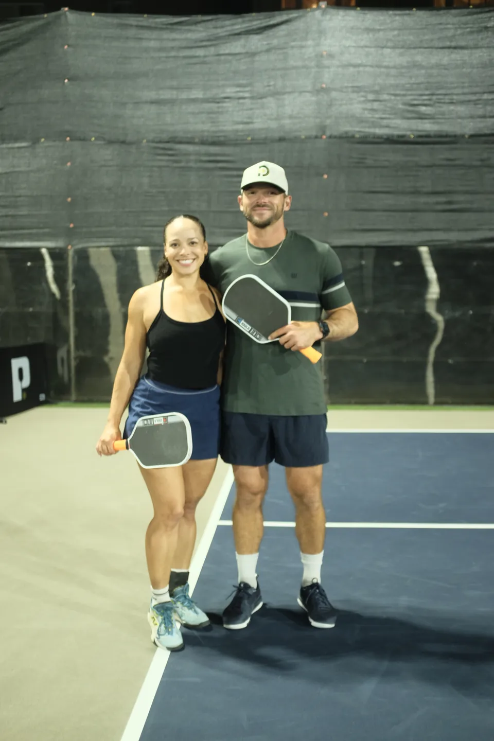 Couple with paddles smile together on court at night, she in a black top and blue skirt, he in a green tee and cap