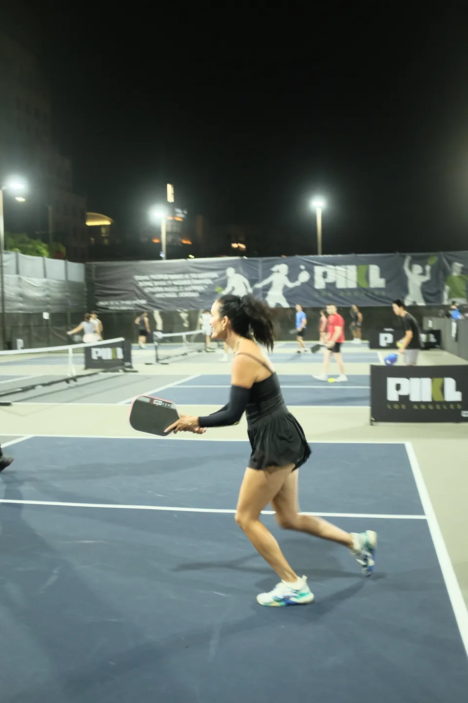 Woman in a black tennis dress and arm sleeve lunges for a shot on a floodlit court, PIKL banners behind